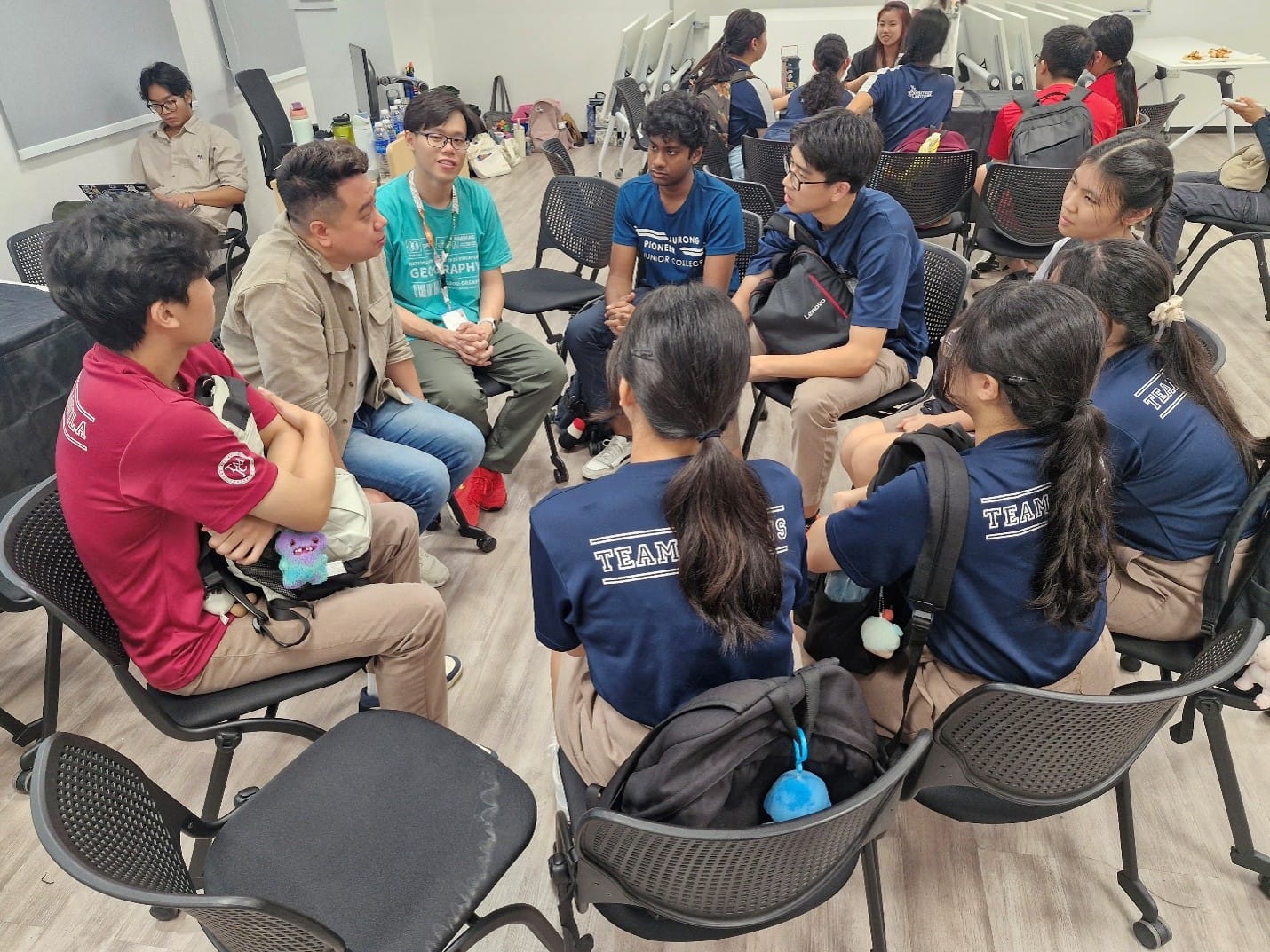 A group of students from different schools sit in a circle of chairs indoors, engaged in a discussion with two adults. The students are wearing various school uniforms.